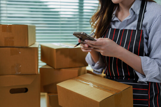Woman standing in apron handling cardboard boxes, checking inventory and order details on a mobile phone for e commerce business