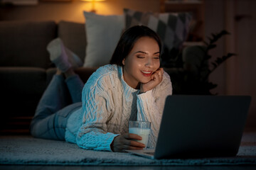 Young woman with glass of milk using laptop at home late in evening