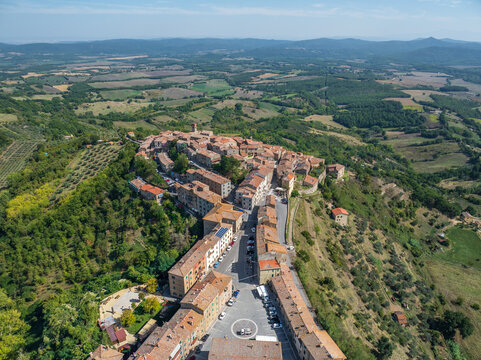 Aerial view of the medieval village perched atop a hill, terracotta roofs contrasting with lush green trees and sunlit walls, Chiusdino, Tuscany, Italy.