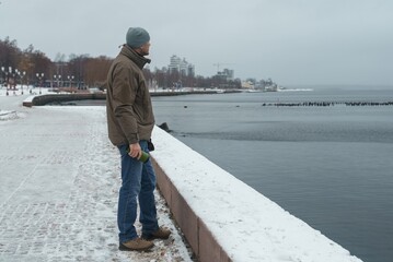 A man stands on the embankment in winter and looks out over the lake. A winter walk along the city's embankment. Relaxing by the lake in winter.