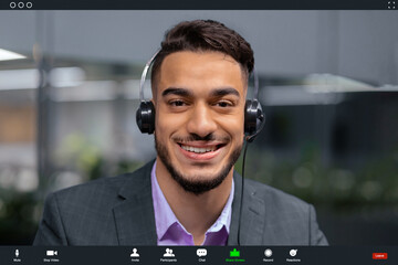 A man with a headset smiles at the camera during a video call in an office environment. He wears a suit and appears engaged in a conversation. The background shows an office space.