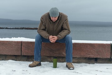 A young man sits in contemplation on the lakeside embankment. The man enjoys a winter rest by the waterside.