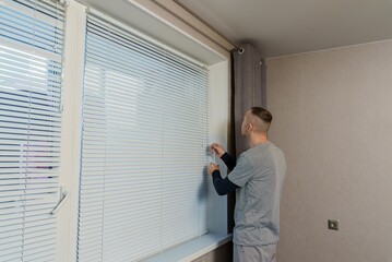 A man is closing the window from daylight with blinds. A man is adjusting the position of the blinds on the window.