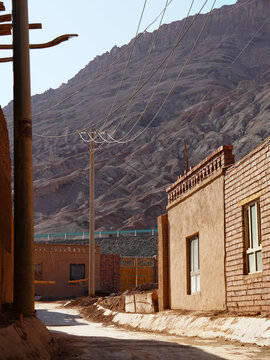 View of sun-baked clay buildings line a narrow street leading towards imposing mountains under a clear sky, creating a stark contrast of textures, Turpan, Xinjiang, China.