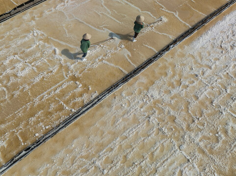 Aerial view of two figures in conical hats traversing the textured salt flats, a mosaic of light and shadow under the Vietnamese sun, Phuoc Diem, Ninh Thuan, Vietnam.