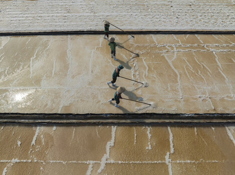 Aerial view of workers meticulously raking salt crystals across the vast, sun-drenched salt pans, creating a mesmerizing pattern of textures and tones, Phuoc Diem, Ninh Thuan, Vietnam.