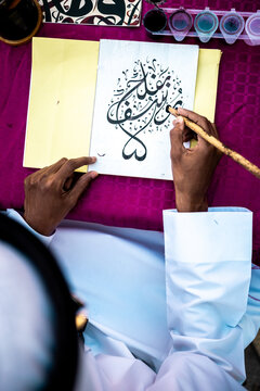 View of a man with an ink pen creating swirling Arabic calligraphy on bright white paper, contrasted against a vibrant magenta surface., Location Omitted, Location Omitted.