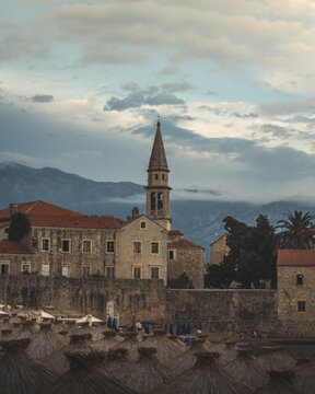 View of old stone buildings with red tile roofs and a tall church steeple rising against a backdrop of hazy mountains, Budva, Budva Municipality, Montenegro.