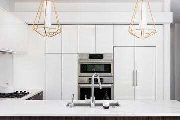 A chrome faucet detail in a white modern kitchen with gold light fixtures hanging over a marble...