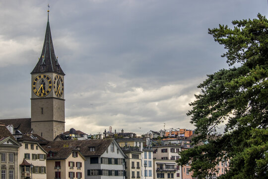 View of the spire of St Peter's church rises above buildings with its golden clock face against a cloudy sky, Zurich, Switzerland.