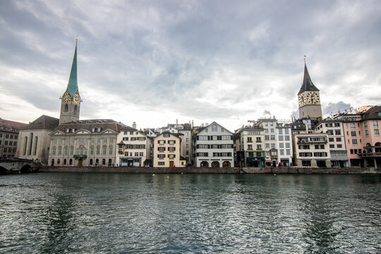 View of buildings with colorful facades and striking towers reflecting in the tranquil river under a cloudy sky, Zurich, Switzerland.