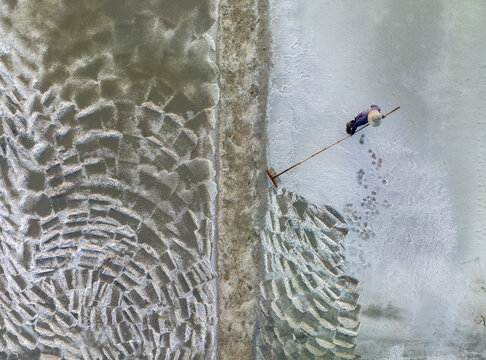 Aerial view of a worker raking through salt fields, the crystalline patterns shimmering under the sun, creating an abstract mosaic, Long Dien, Ba Rịa - Vung Tau, Vietnam.