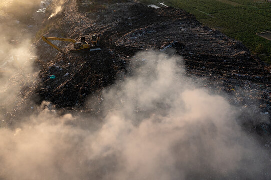 Aerial view of a vast landfill where a yellow excavator toils amidst the dark waste and rising smoke, creating a scene of stark contrast, Chattogram, Chittagong Division, Bangladesh.
