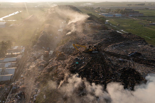 Aerial view of bulldozers atop a massive landfill, smoke rising against a backdrop of green fields and distant buildings, Chattogram, Chittagong Division, Bangladesh.