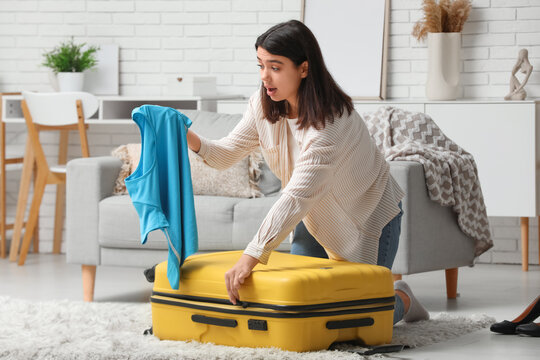 Surprised woman packing blue t-shirt into suitcase with clothes in living room - Powered by Adobe