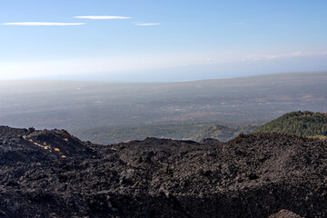 Obraz premium Volcanic landscape on the slopes of Mount Etna, Sicily, Italy. Dark solidified lava fields in the foreground with a panoramic view over the Sicilian countryside and distant coastline under a clear sky
