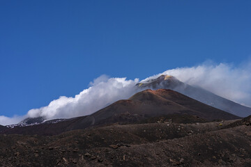 Summit of Mount Etna volcano rising above dark volcanic slopes, partially wrapped in white clouds under a clear blue sky. The image showcases the dramatic volcanic landscape of Sicily, Italy