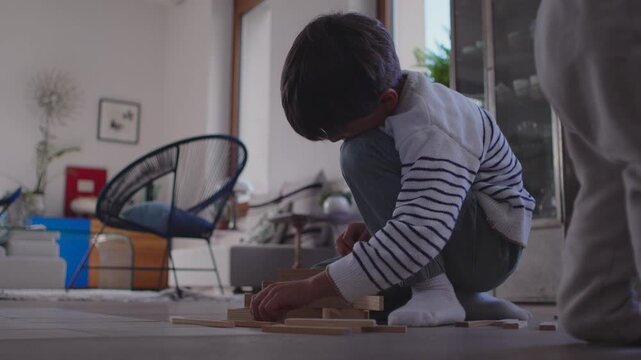 Boy assembling wooden blocks on floor with calm steady focus while carefully stacking pieces during slow thoughtful playtime creating peaceful moment of concentration in cozy home setting