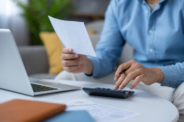 Man holding a paper document and using a calculator while sitting at a table with a laptop,...