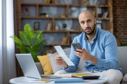 Male freelancer sitting on a couch at home, looking stressed while holding a long bill and using his smartphone for mobile banking or budgeting, managing personal finances with a laptop nearby - Powered by Adobe