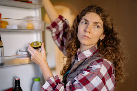 A woman with curly hair looks thoughtfully at an avocado as she stands in front of an open refrigerator in a kitchen. She seems to decide on a meal