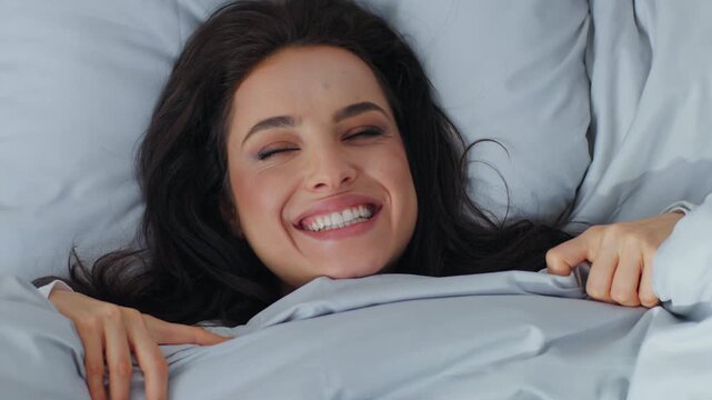 Close up playful Caucasian woman peeking out hiding under blanket duvet laughing sincere looking at camera smiling toothy laugh flirting girl female waking up in good morning awaking lying in bed