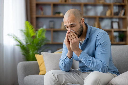 Young man using a tissue to blow his nose while sitting on a couch at home, experiencing symptoms of a cold, flu, virus, or seasonal allergy and needing relief