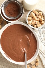 Liquid chocolate dough in bowl, cocoa powder, brown sugar and milk on wooden table, flat lay
