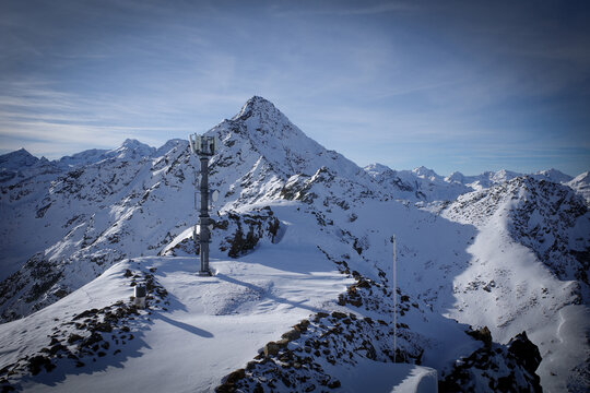 A striking winter mountain landscape featuring a telecommunications tower standing on a snow-covered alpine ridge with dramatic peaks in the background