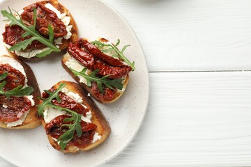 Delicious bruschettas with cream cheese, sun-dried tomatoes and arugula on white wooden table, top view. Space for text
