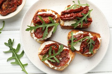 Delicious bruschettas with cream cheese, sun-dried tomatoes and arugula on white wooden table, flat lay