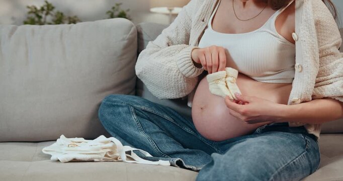 Pregnant woman sitting on couch folding tiny baby clothes and preparing for newborn arrival with anticipation and care. Pregnant Woman Preparing Baby Clothes