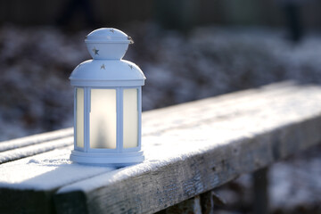 White lantern with star designs sits on a snow-covered wooden bench, casting a warm glow in a serene winter landscape, evoking feelings of peace and tranquility