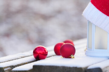 Red Christmas ornaments scattered on a snowy wooden surface beside a lantern with a cozy knitted hat, creating a festive winter atmosphere with holiday spirit
