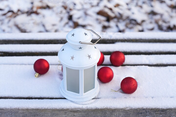 White lantern with star designs sits on a snowy wooden surface surrounded by red Christmas ornaments, creating a festive winter atmosphere with holiday charm
