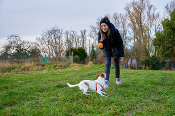 Woman standing on grass holding ball while small dog runs toward her