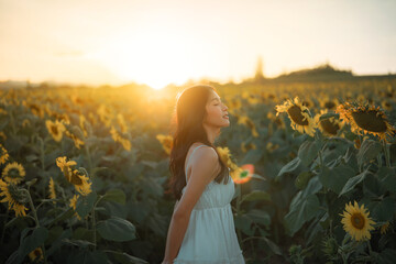 Happy young asian woman standing in beautiful sunflower field during sunset or sunrise with sun flare for summer travel and freedom nature concept