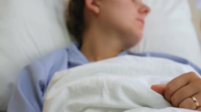 Close up focus pull from an intravenous catheter in a young woman's hand to her face as she rests in a hospital bed, receiving medical treatment and looking away with a thoughtful expression