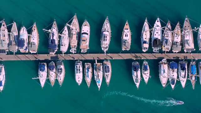Sailboats docked in orderly rows large marina green water many ships. Drone overhead shot of sailboats neatly docked along symmetrical wooden pier Greece