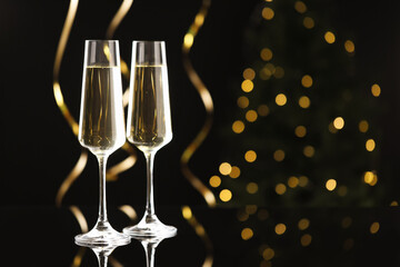 Merry Christmas and Happy New Year. Flute glasses with sparkling wine and serpentine streamers on black background against blurred lights. Space for text