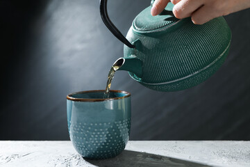 Woman pouring hot tea into cup from teapot at grey table, closeup