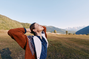 Fototapeta premium Woman in nature among mountains and a wide field, enjoying relaxation and freedom outdoors during a travel break, eyes closed and arms raised in a peaceful autumn landscape.
