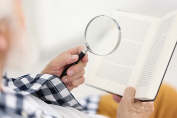 Senior man using magnifying glass while reading book indoors, closeup