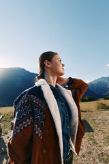 Fototapeta premium Woman in mountains wearing a warm jacket outdoors, portrait in nature with sunlight on face and meadow background, young traveler with braided hair enjoying scenic landscape.