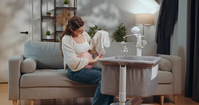 Expecting mother sits on sofa holding baby clothes near bedside bassinet with mobile. Pregnant Woman Preparing Baby Crib