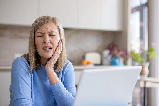 Senior woman clutching her jaw in pain while seated with a laptop at home, grimacing from severe toothache and urgently needing dental care and relief