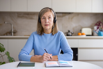 Naklejka premium Mature woman in her kitchen wearing a headset and holding a pen, focused on a laptop during a video conference or e-learning session, taking notes and working from home