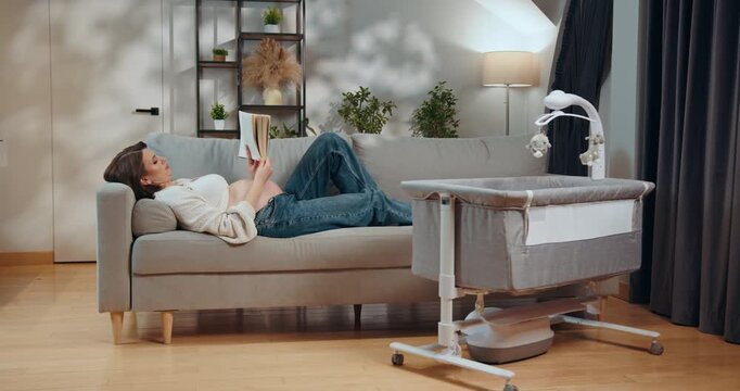 Pregnant woman rests on sofa reading book near baby crib preparing for motherhood and new life. Pregnant Woman Reading on Sofa