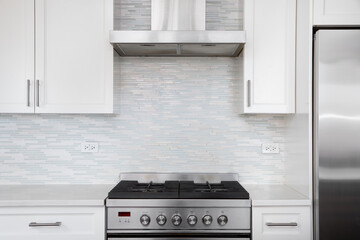 Fototapeta premium A modern white kitchen features a stainless steel gas stove against a light blue and white mosaic tile backsplash. 