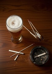 An overhead view of a wooden table holding a glass of frothy beer, an ashtray, and several tobacco sticks for smoking ,filter ,lager ,nicotine